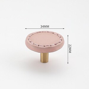 May include: A pink leather cabinet knob with a stitched edge and a brass base. The knob measures 34 mm in diameter and 22 mm in height.