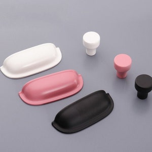 Może przedstawiać: Cztery gałki do szafki i trzy uchwyty do szuflad w kolorze czarnym, białym i różowym. Gałki są okrągłe, a uchwyty prostokątne.