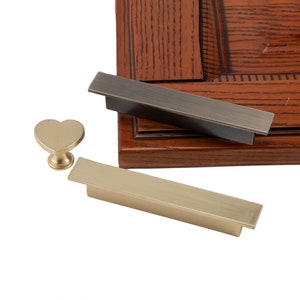 May include: Two gold cabinet pulls and a heart-shaped knob. The pulls are rectangular and have a brushed finish. The knob is a simple heart shape.