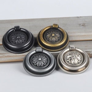 Puede incluir: Cuatro pomos decorativos para armario con tirador de anillo. Los pomos son de acabado negro, oro antiguo, plata y gris. Cada pomo presenta un diseño floral en relieve.