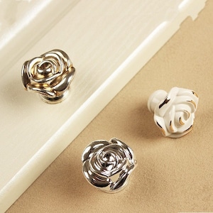 Può includere: Primo piano di tre pomelli per mobili a forma di rosa. Due sono color argento e uno bianco con dettagli dorati. I pomelli sono probabilmente in metallo e ceramica. Questi pomelli decorativi sono progettati per mobili.