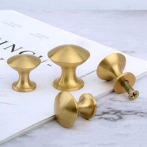 Op de afbeelding: Drie gouden kastknoppen met een ronde, licht gewelfde bovenkant. De knoppen zijn van verschillende grootte en hebben een gladde, gepolijste afwerking.