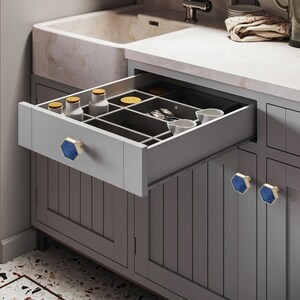 Pu&ograve; includere: Un mobile da cucina grigio con un cassetto aperto, che rivela un organizzatore grigio con scomparti. Il cassetto ha una maniglia esagonale blu e dorata. Il mobile ha tre maniglie esagonali blu e dorate.