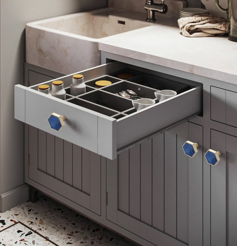 Pu&ograve; includere: Un mobile da cucina grigio con un cassetto aperto, che rivela un organizzatore grigio con scomparti. Il cassetto ha una maniglia esagonale blu e dorata. Il mobile ha tre maniglie esagonali blu e dorate.