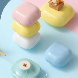 May include: A selection of colourful cabinet knobs in pink, yellow, white, and light blue. One yellow knob features a small cat figurine. The knobs have a square, rounded design, suitable for drawers or cupboards.