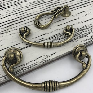 Puede incluir: Tres tiradores de armario de bronce antiguo con diseños ornamentados. Dos son tiradores curvos con un anillo decorativo en el centro. El tercero es un tirador de anillo con un adorno decorativo en la parte superior.