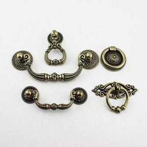 Może przedstawiać: Zestaw sześciu antycznych gałek i uchwytów do szafek w wykończeniu mosiężnym. Gałki mają ozdobne wzory z uchwytem w kształcie pierścienia i okrągłą gałką z kwiatowym wzorem. Uchwyty mają zakrzywiony kształt z ozdobnym wzorem.