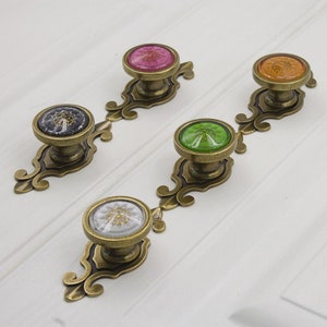 Puede incluir: Cinco pomos de armario de estilo antiguo con bases de metal ornamentadas e inserciones de vidrio de colores. Los pomos están dispuestos en una fila sobre una superficie blanca.