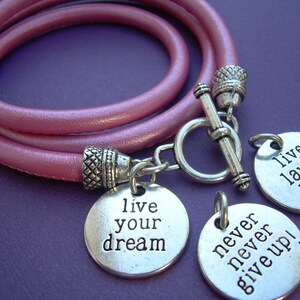 Op de afbeelding: Roze leren wikkelarmband met zilverkleurige bedels. De bedels hebben de tekst "live your dream", "never never give up!" en "live laugh love".