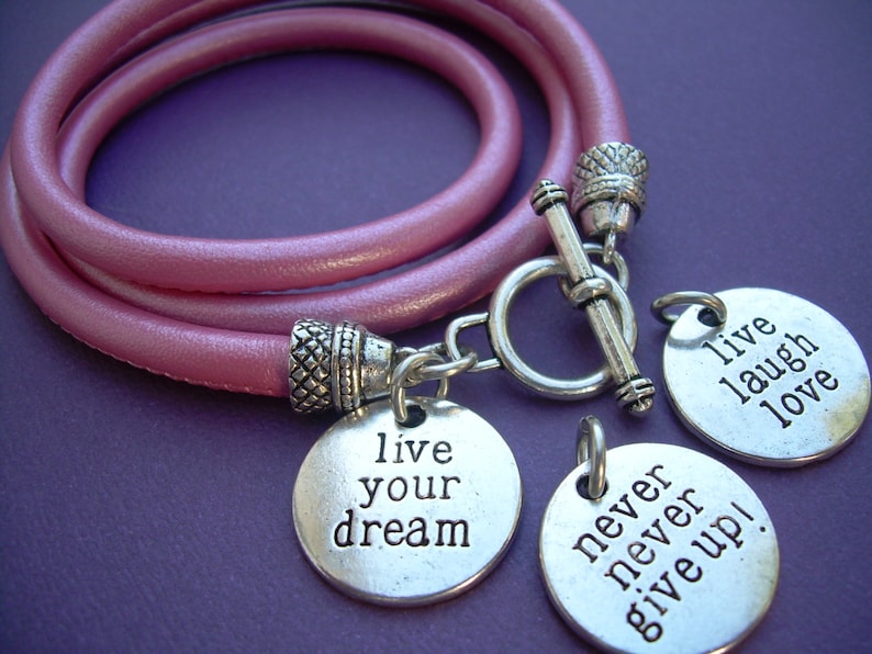 Op de afbeelding: Roze leren wikkelarmband met zilverkleurige bedels. De bedels hebben de tekst "live your dream", "never never give up!" en "live laugh love".
