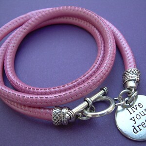 Op de afbeelding: Een roze leren armband met een zilveren bedel met de tekst "live your dream".