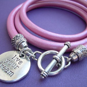 Op de afbeelding: Een roze lederen armband met een zilveren schuifgesp en een zilveren bedel met de tekst "Live your dream".