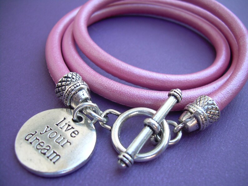 Op de afbeelding: Een roze lederen armband met een zilveren schuifgesp en een zilveren bedel met de tekst "Live your dream".
