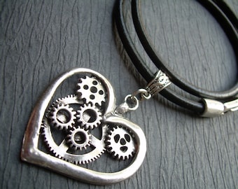 May include: A silver heart-shaped pendant necklace with a design of interlocking gears. The necklace is on a black leather cord.
