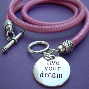 Op de afbeelding: Een roze leren armband met een zilveren bedel met de tekst "Live your dream".