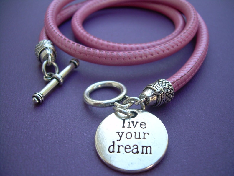 Op de afbeelding: Een roze leren armband met een zilveren bedel met de tekst "Live your dream".