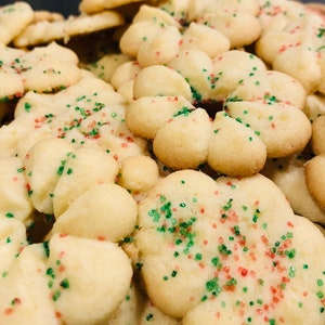 May include: Close-up of a pile of flower-shaped cookies with green and red sprinkles. The cookies are a light golden color and appear to be freshly baked. The image is well-lit, showcasing the texture and details of the treats.