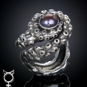 May include: A silver ring with an intricate design resembling an octopus tentacle. The ring features a large, dark purple pearl in the centre.