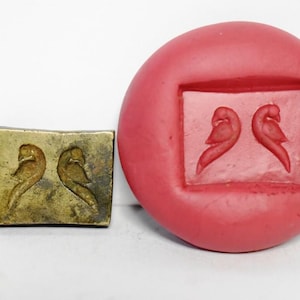 Può includere: Un timbro vintage in ottone e uno stampo in gomma rossa, entrambi con un disegno di due uccelli stilizzati rivolti l'uno verso l'altro. Il timbro in ottone è rettangolare, mentre lo stampo è rotondo. Gli uccelli sono raffigurati in uno stile semplice ed elegante.