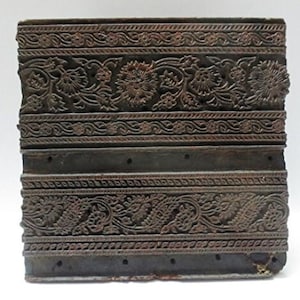 May include: A dark brown wooden printing block with two rows of intricate floral designs carved into the surface. The block has a worn, aged appearance and is likely used for textile printing.