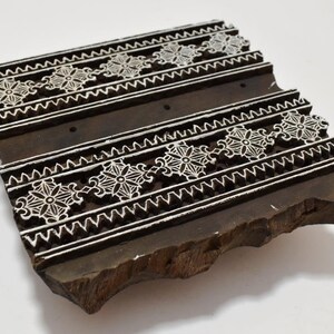 May include: A wooden printing block with a carved design of white floral patterns on a dark brown background. The block is rectangular and has a rough, uneven surface.