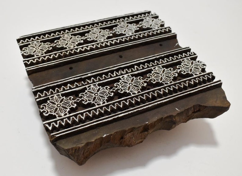 May include: A wooden printing block with a carved design of white floral patterns on a dark brown background. The block is rectangular and has a rough, uneven surface.