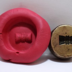 Könnte beinhalten: Ein roter Tonabdruck eines Stempels aus Messing mit einem Schleifendesign. Der Messingstempel ist ebenfalls im Bild zu sehen.