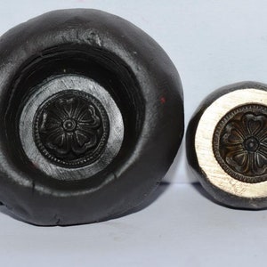 Könnte beinhalten: Eine schwarze Gummiform und eine Messingform mit einem floralen Design. Beide Formen sind rund und haben ein erhabenes Design in der Mitte.