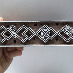 May include: A wooden printing block featuring a repeating pattern of white geometric shapes and swirls. The block is brown and has a rough texture.