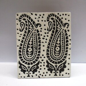 Puede incluir: Un bloque de impresión de madera blanco con dos diseños paisley negros. El bloque tiene un patrón de puntos alrededor de los diseños paisley.