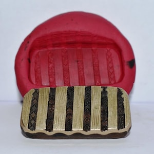 Peut inclure: Gros plan d'un moule en caoutchouc rouge avec un motif décoratif. Un tampon en laiton avec un motif similaire est placé devant le moule.