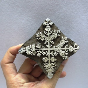 Könnte beinhalten: Ein quadratischer Holzblock-Stempel mit einem floralen Design. Das Design besteht aus mehreren weißen floralen Motiven auf einem dunkelbraunen Hintergrund.