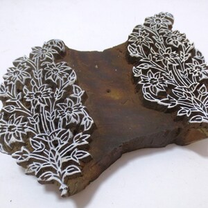 May include: A wooden printing block featuring an intricate floral design. The block is carved with delicate details of flowers and leaves. The design is painted white on the brown wood.