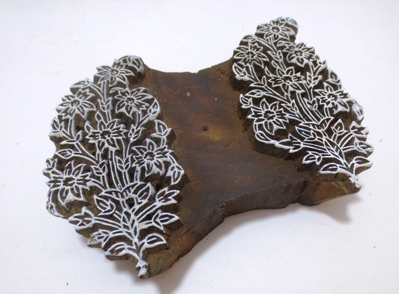 May include: A wooden printing block featuring an intricate floral design. The block is carved with delicate details of flowers and leaves. The design is painted white on the brown wood.