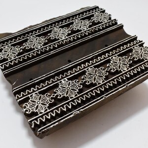 May include: A wooden printing block with a carved design of white floral patterns on a dark brown background. The block is rectangular and has a slightly uneven surface.