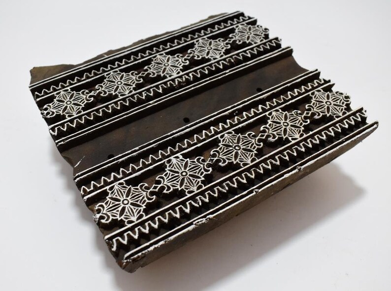 May include: A wooden printing block with a carved design of white floral patterns on a dark brown background. The block is rectangular and has a slightly uneven surface.