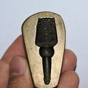 May include: A brass mold for casting a decorative object. The mold is shaped like a teardrop and has a detailed design of a stylized figure with a crown and a long, pointed hat.