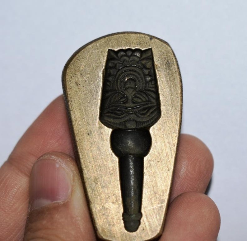 May include: A brass mold for casting a decorative object. The mold is shaped like a teardrop and has a detailed design of a stylized figure with a crown and a long, pointed hat.