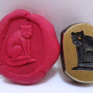 May include: A close-up of a brass stamp with a black cat design. The stamp is used to create an impression in red clay, which is also shown in the image.