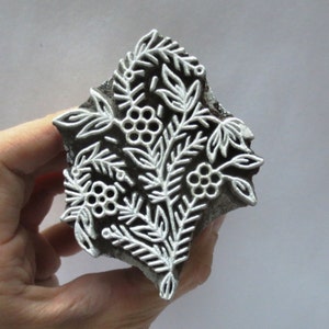 May include: A dark grey wooden block with a white floral pattern. The design features leaves and berry-like clusters. The block is held in a hand.