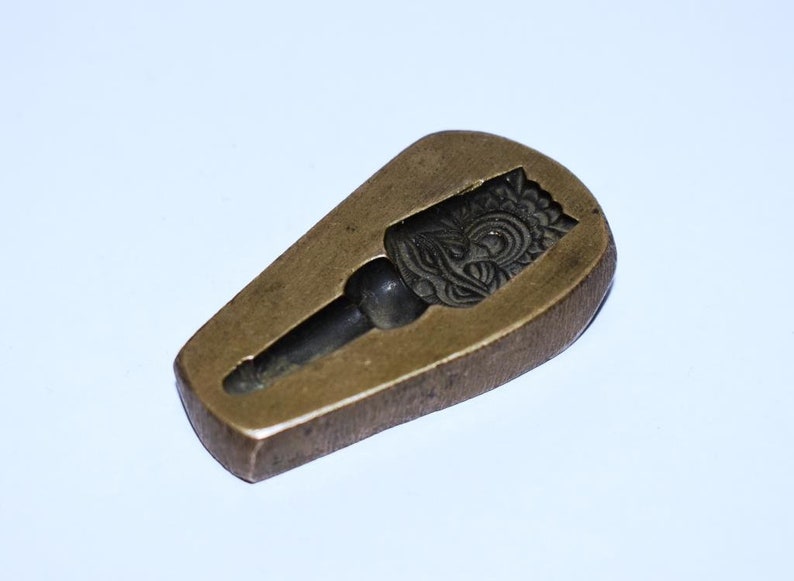 May include: A vintage brass mold for making a decorative object. The mold features a detailed design of a face with intricate patterns.