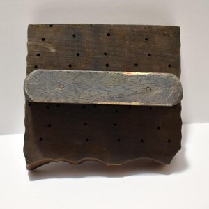 May include: A wooden printing block with a handle. The block has a grid of holes and a rough, uneven edge.