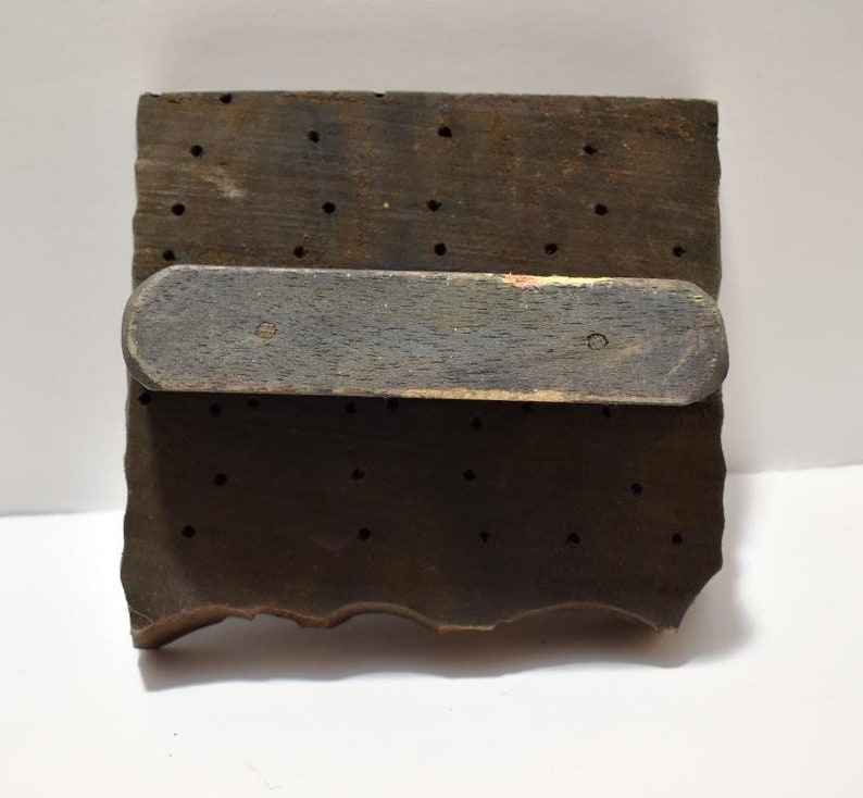 May include: A wooden printing block with a handle. The block has a grid of holes and a rough, uneven edge.
