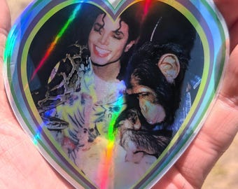 May include: A heart-shaped sticker featuring a photographic print of Michael Jackson smiling next to a chimpanzee. The sticker has a holographic border with concentric bands of green, blue, and silver that reflect light in a rainbow spectrum.