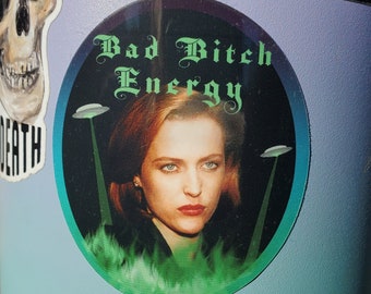 May include: A round sticker with a black background and a green glow around the edges. The sticker features a woman with brown hair and a green background. The text on the sticker reads "Bad Bitch Energy".