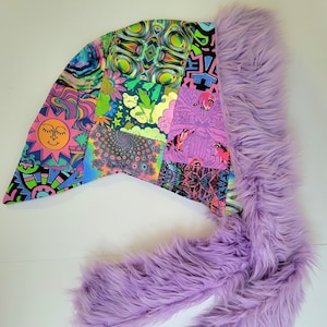 Może przedstawiać: Kaptur w stylu patchwork z żywym, psychodelicznym wzorem i długim, puszystym, lawendowym wykończeniem z imitacji futra. Kaptur posiada różnorodne wzory, w tym uśmiechnięte słońce, kwiaty i kształty geometryczne. Wykończenie z imitacji futra dodaje miękki, dotykowy element.