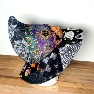 May include: A bonnet featuring a patchwork design with various celestial and mystical patterns. The fabric includes black and white spiderwebs, a purple and gold sun and moon print, black cat faces, geometric eye motifs, and abstract star patterns.