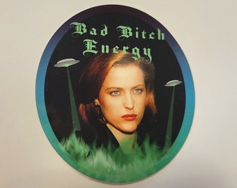 May include: A round sticker with a dark blue background and a green and blue gradient around the edges. The sticker features a woman with brown hair and a black jacket. The text "Bad Bitch Energy" is written in a gothic font in green on the sticker. Two flying saucers with green beams are in the background.