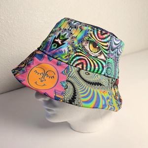 May include: A colorful bucket hat with a patchwork design. The hat features a pink panel with a smiling sun, and other panels with psychedelic patterns, including an eye and swirling designs. The hat is displayed on a white mannequin head.