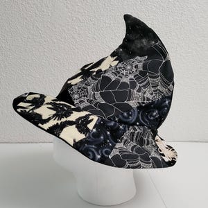 Pode incluir: Um chapéu de bruxa preto e branco com uma coroa pontuda e aba larga. O chapéu apresenta um design de patchwork com padrões de corvos, teias de aranha e celestes. O chapéu é exibido em uma cabeça de manequim branca.